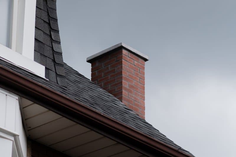 Dark asphalt shingles and a brick chimney on a brick home under an overcast sky